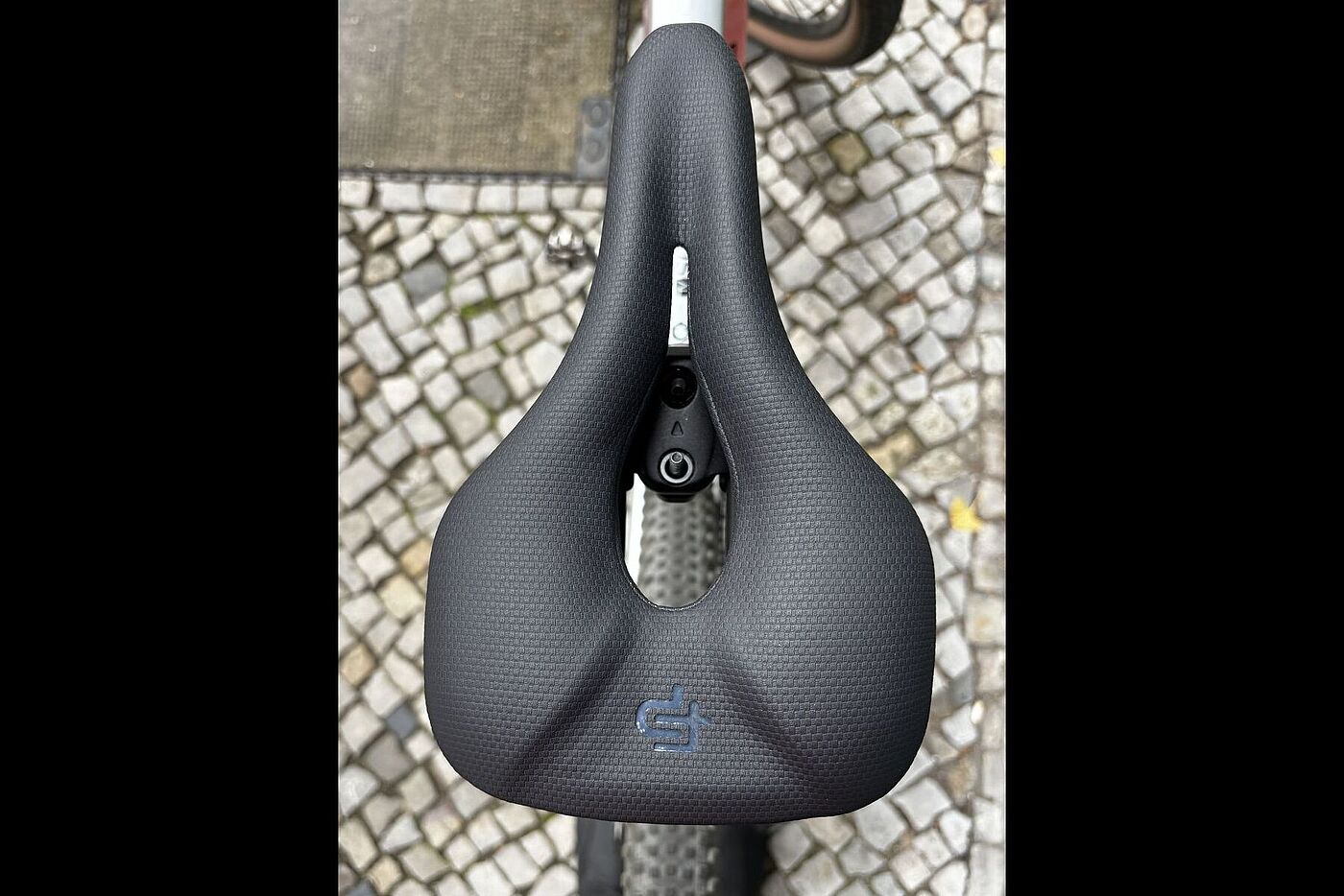 Schwarzer Fahrradsattel mit großer mittlerer Öffnung, von oben auf Kopfsteinpflaster fotografiert.