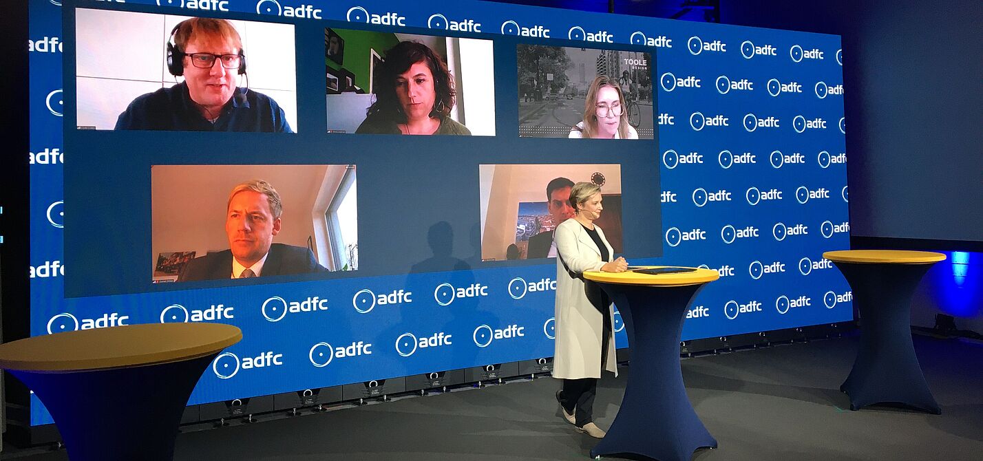 Symposium2_Copyright_Christin_Stein Das digitale ADFC-Symposium wurde aus einem Studio mit zugeschalteten Expert*innen übertragen. Moderatorin Janine Steeger (vorne) führte durch das Programm. Im Hintergund zu sehen: Oben von links nach rechts Clyde Loakes (London), Silvia Casorrán (Barcelona), Jennifer Toole (Maryland). Unten von links: Daniel Zöhler (Bocholt), Martin Hikel (Berlin, verdeckt)