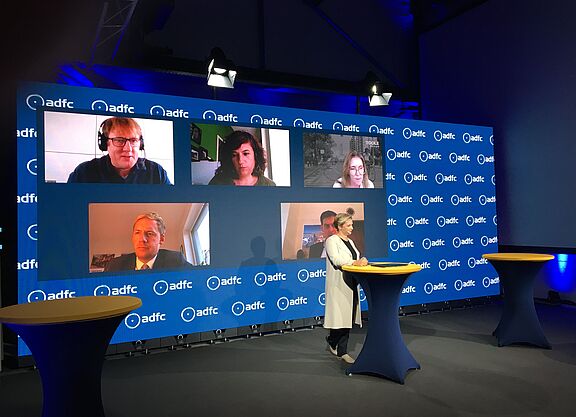 Symposium2_Copyright_Christin_Stein Das digitale ADFC-Symposium wurde aus einem Studio mit zugeschalteten Expert*innen übertragen. Moderatorin Janine Steeger (vorne) führte durch das Programm. Im Hintergund zu sehen: Oben von links nach rechts Clyde Loakes (London), Silvia Casorrán (Barcelona), Jennifer Toole (Maryland). Unten von links: Daniel Zöhler (Bocholt), Martin Hikel (Berlin, verdeckt)