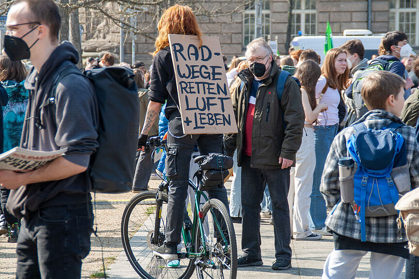 Demonstration im Freien. Im Vordergrund steht eine Person mit einem Fahrrad, die ein Schild mit der Aufschrift „Radwege retten Luft und Leben trägt“